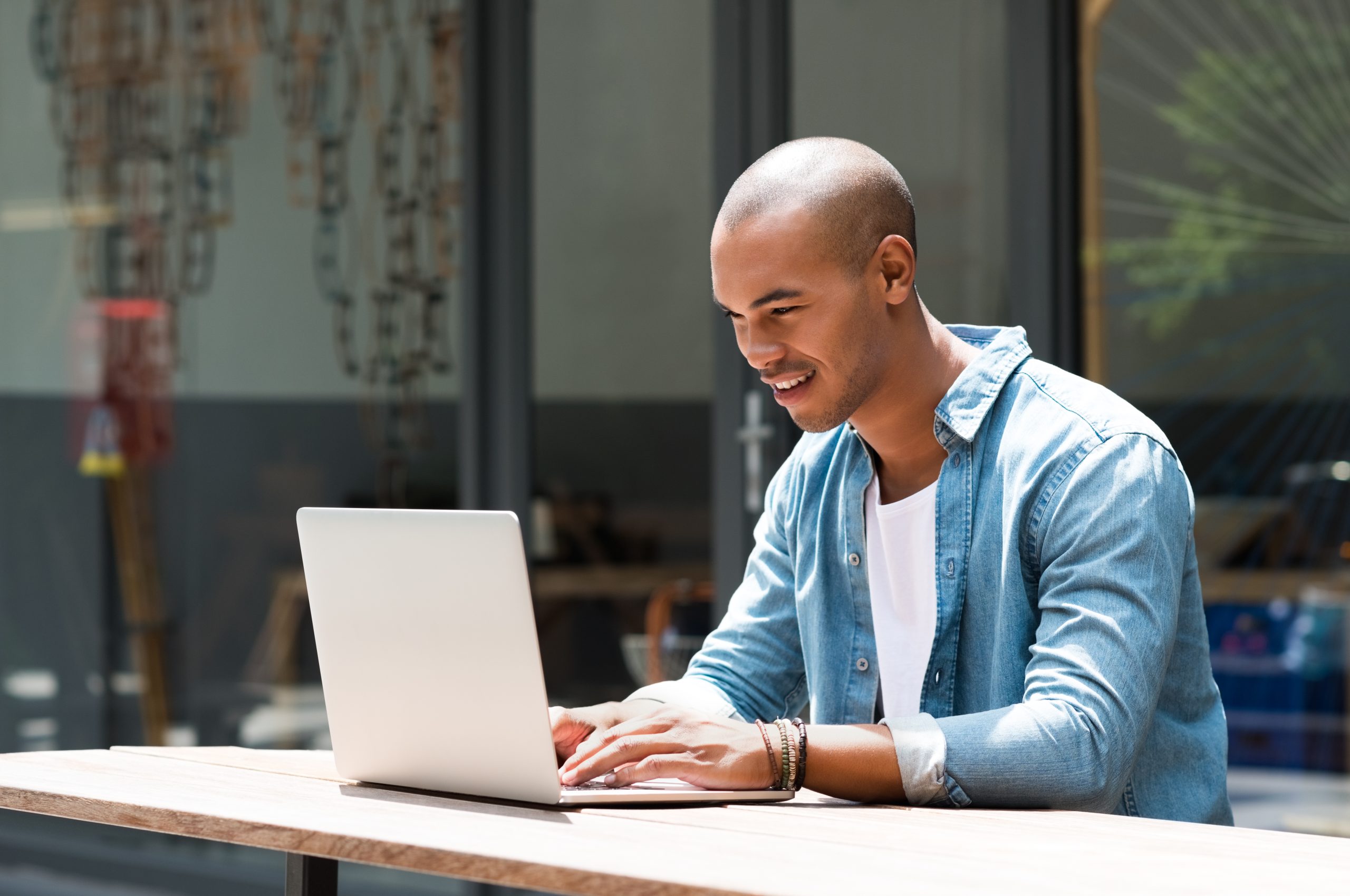 Happy man writing text on laptop in a modern coffee shop. Happy african student surfing the web on laptop with wifi free connection. Smiling guy in casual studying on laptop sitting outdoor the cafe.